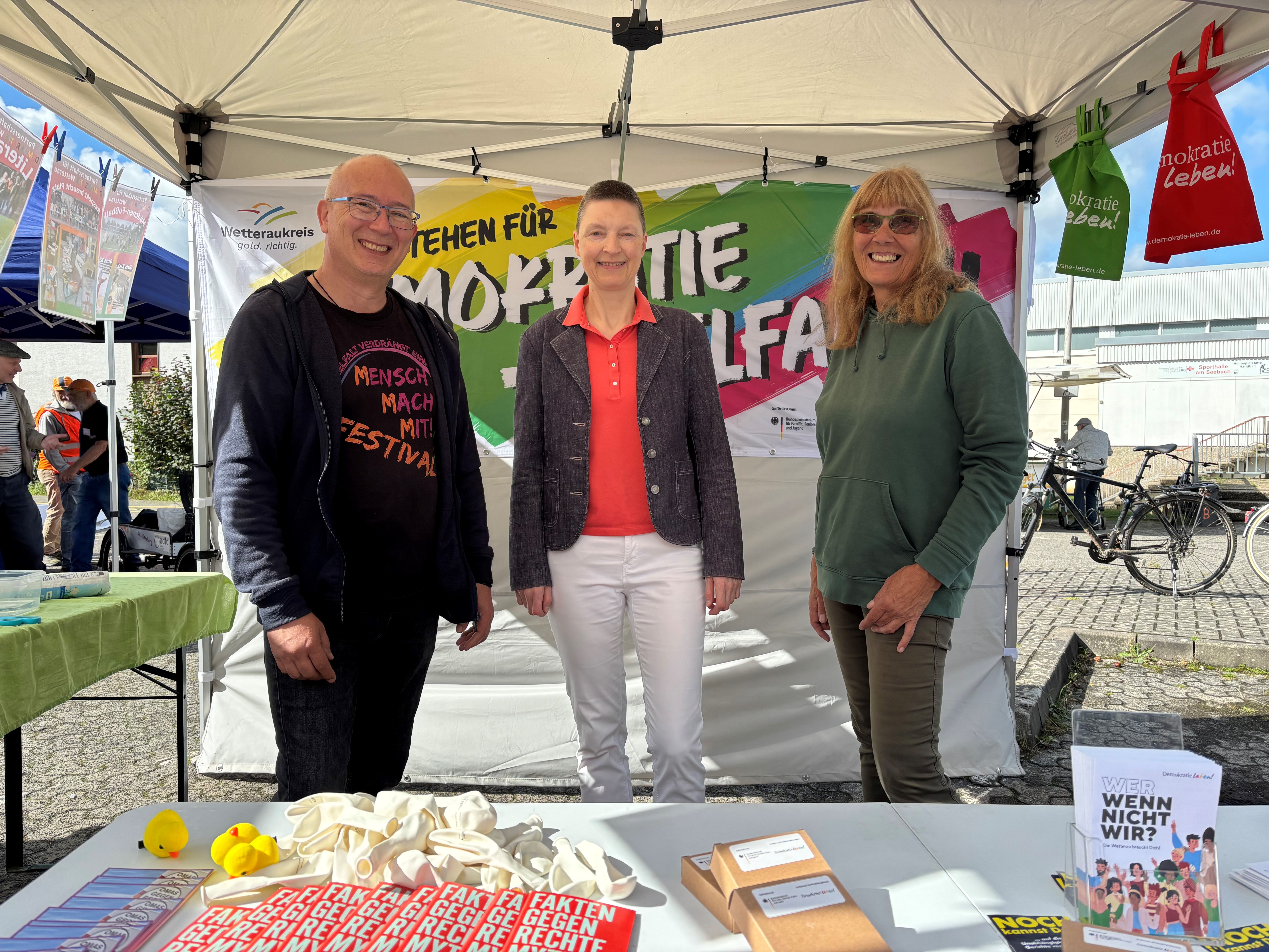 Zwei Frauen und ein Mann stehen gemeinsam an einem Infostand.