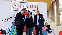 Eine Frau und zwei Männer stehen zusammen in einem Säulengang vor einer großen, bunten Plakatwand mit der Aufschrift "Frühe Hilfen".