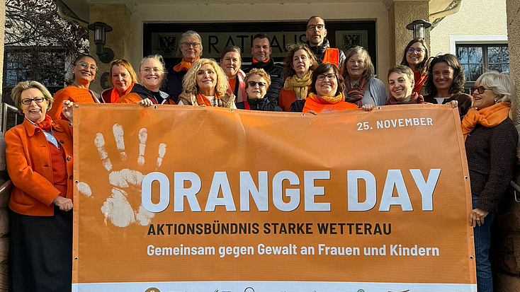Eine Menschengruppe steht zusammen auf einer Treppe und zeigt ein großes Plakat. Auf diesem steht die Aufschrift "Orange Day Aktionsbündnis Starke Wetterau, Gemeinsam gegen Gewalt an Frauen und Kindern.