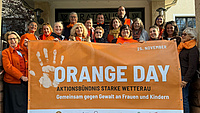 Eine Menschengruppe steht zusammen auf einer Treppe und zeigt ein großes Plakat. Auf diesem steht die Aufschrift "Orange Day Aktionsbündnis Starke Wetterau, Gemeinsam gegen Gewalt an Frauen und Kindern.