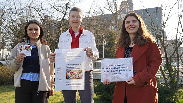 Drei Frauen stehen im Freien auf einem Platz, im Hintergrund sind Büsche und Gebäude zu sehen. Zwei der Frauen halten Plakate in Hände, die dritte mehrere Karten.