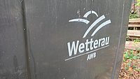 Eine Mülltonne mit der Aufschrift AWB und einem Logo steht im Freien.
