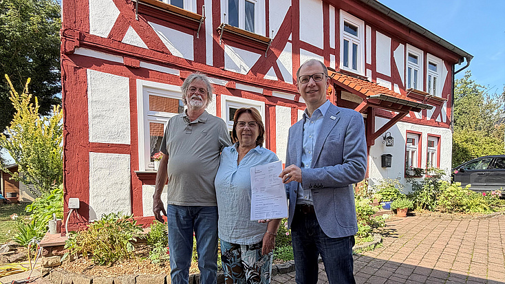Eine Frau und zwei Männer stehen auf einem Hof vor einem Fachwerkgebäude. Einer der Männer hält ein Dokument in Händen.