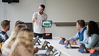 Mehrere Menschen sitzen um einen Tisch herum, einer steht vorne und erklärt etwas.