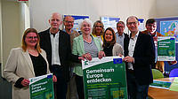 Vier Frauen und fünf Männer stehen in einem Raum. Mehrere von ihnen halten Plakate in den Händen.