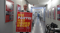 Ein Mensch steht vor einer langen Wand, er hält ein Plakat in den Händen.