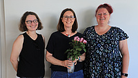 Drei Frauen stehen in einem Raum vor einer Wand, eine von ihnen hält einen Blumenstrauß in den Händen.