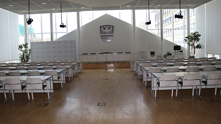 In einem großen Saal stehen Stuhl- und Tischreihen in Richtung eines Podests und Rednerpults ausgerichtet. An der Stirnseite des Saal befindet sich eine Fensterfront, und ein großes Wappen hängt an der Wand.