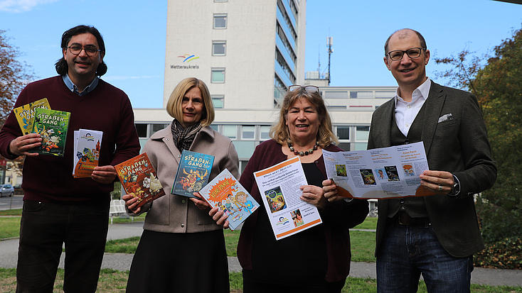Zwei Frauen und zwei Männer stehen auf einem Platz im Freien, im Hintergrund ist ein Hochhaus zu sehen. Alle vier halten Bücher und Flyer in Händen.