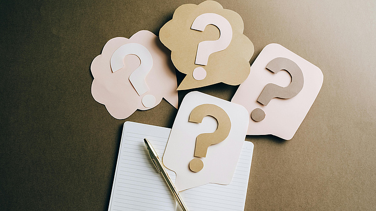 Auf einer Tischoberfläche liegen ein Notizbuch, ein Stift und vier grafische Elemente, die Fragezeichen in Sprechblasen darstellen.