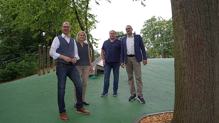 Vier Personen stehen auf einer neu gestalteten Spielfläche an einem Hang. Der versickerungsfähige Belag ist grün, im Hintergrund ist eine Rutsche zu sehen.