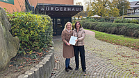 Zwei Frauen stehen vor einem Bürgerhaus und übergeben eine Mappe mit einem Zuwendungsbescheid.