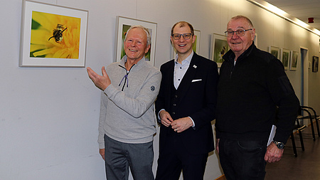 Drei Männer stehen nebeneinander in einem Flur. Der Mann ganz links deutet auf eine Fotografie, die an der Wand hängt.