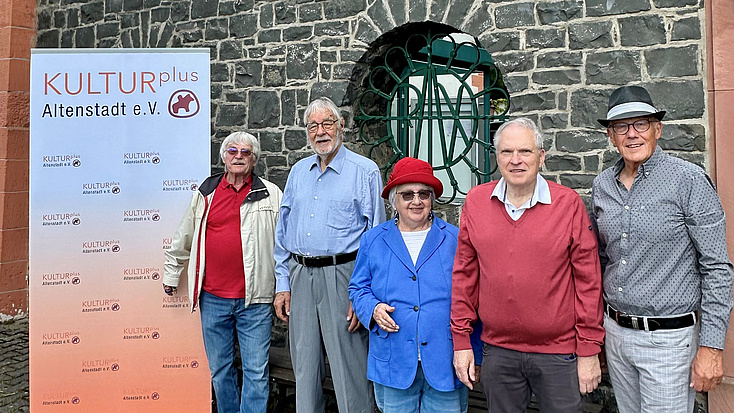 Das Bild zeigt fünf Personen vor einger Steinmauer neben einem Roll-Up des Vereins KULTURplus.