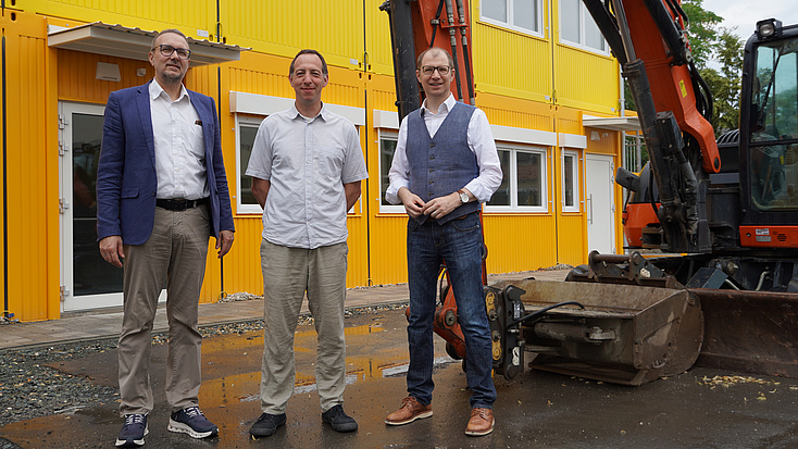 Drei Personen besichtigen die Baumaßnahmen für einen mobiles Raumsystenm. Sie stehen vor dem gelben neuen Räumen für die Haingrabenschule, daneben steht ein Bagger.