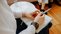 Eine Person sitzt auf einem Stuhl neben einem Tisch und hält einen Stift und ein Notizbuch im Schoß. Ihr gegenüber sitzen weitere Personen.