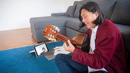 Ein Mann spielt Gitarre. Vor ihm steht ein Tablet auf dem ein Schüler mit Gitarre zu sehen ist.
