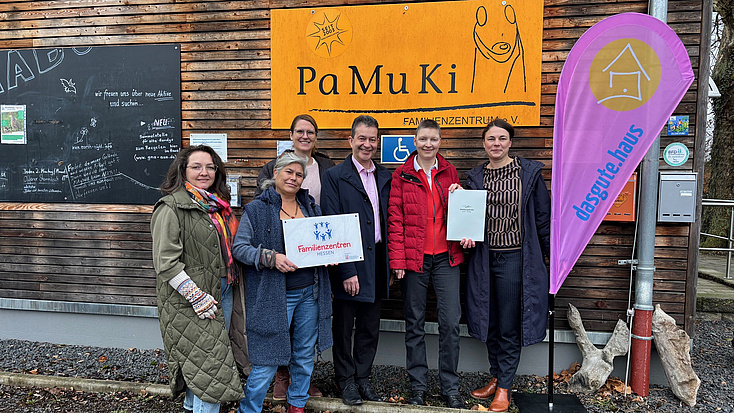 Fünf Frauen und ein Mann stehen vor einer Hauswand. An dieser ist ein Logo zu sehen, ein weiteres findet sich auf einer Beachflag neben der Personengruppe. Eine der Frauen hält eine Tafel mit einem weiteren Logo in Händen, zwei weitere halten gemeinsam ein Dokument hoch.