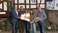 Zwei Frauen und drei Männer stehen gemeinsam vor einem Fachwerkhaus. Eine der Frauen und einer der Männer halten gemeinsam eine Dokumentenmappe in Händen. Ein weiterer Mann hält den Ausdruck einer Internetseite in der Hand.