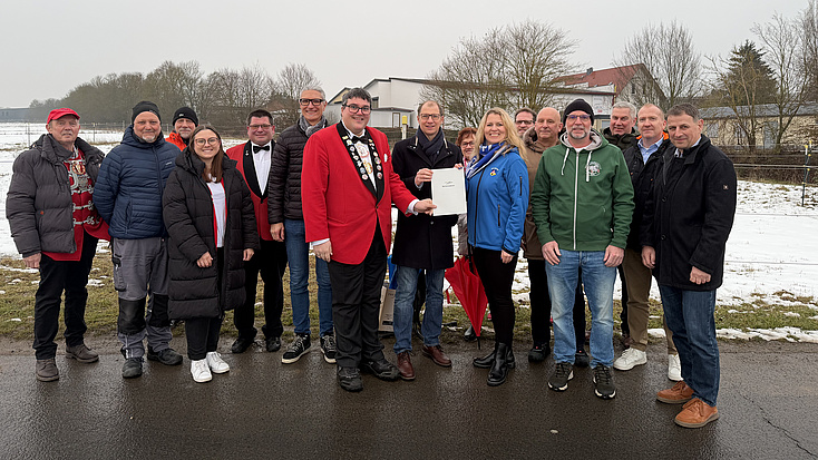 Eine größere Personengruppe steht gemeinsam auf einem Feldweg. Zwei Männer halten gemeinsam ein Dokument in Händen. Im Hintergrund sind eine verschneite Wiese und mehrere Gebäude zu erkennen.